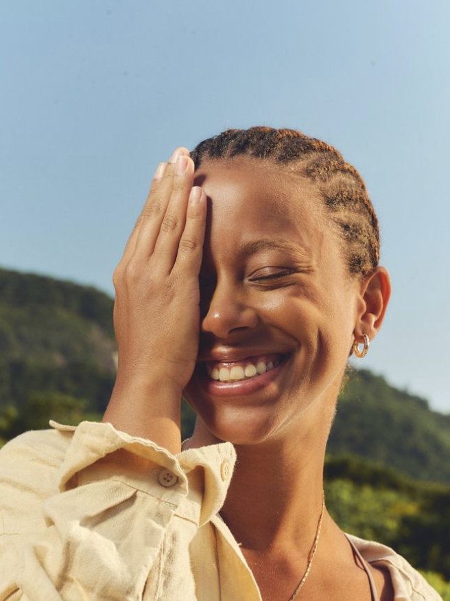 Laughing woman with braided hair in a pale yellow linen shirt, hand over one eye, green hills behind her